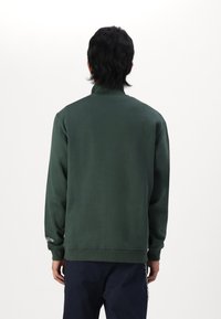Sudadera verde con un cuello alto, puños y dobladillo acanalados. Presenta una textura suave con un detalle en la manga izquierda.