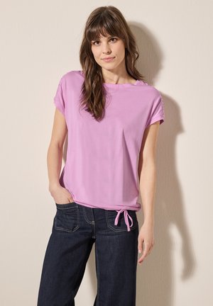 Lichtpaarse t-shirt met korte mouwen, een relaxed fit en zijstrikdetail, gecombineerd met donkerblauwe wijde jeans. Gladde stoftextuur.