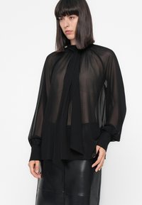 Donna che indossa una blusa nera trasparente a maniche lunghe con un nodo al collo e una gonna di pelle nera su uno sfondo bianco.