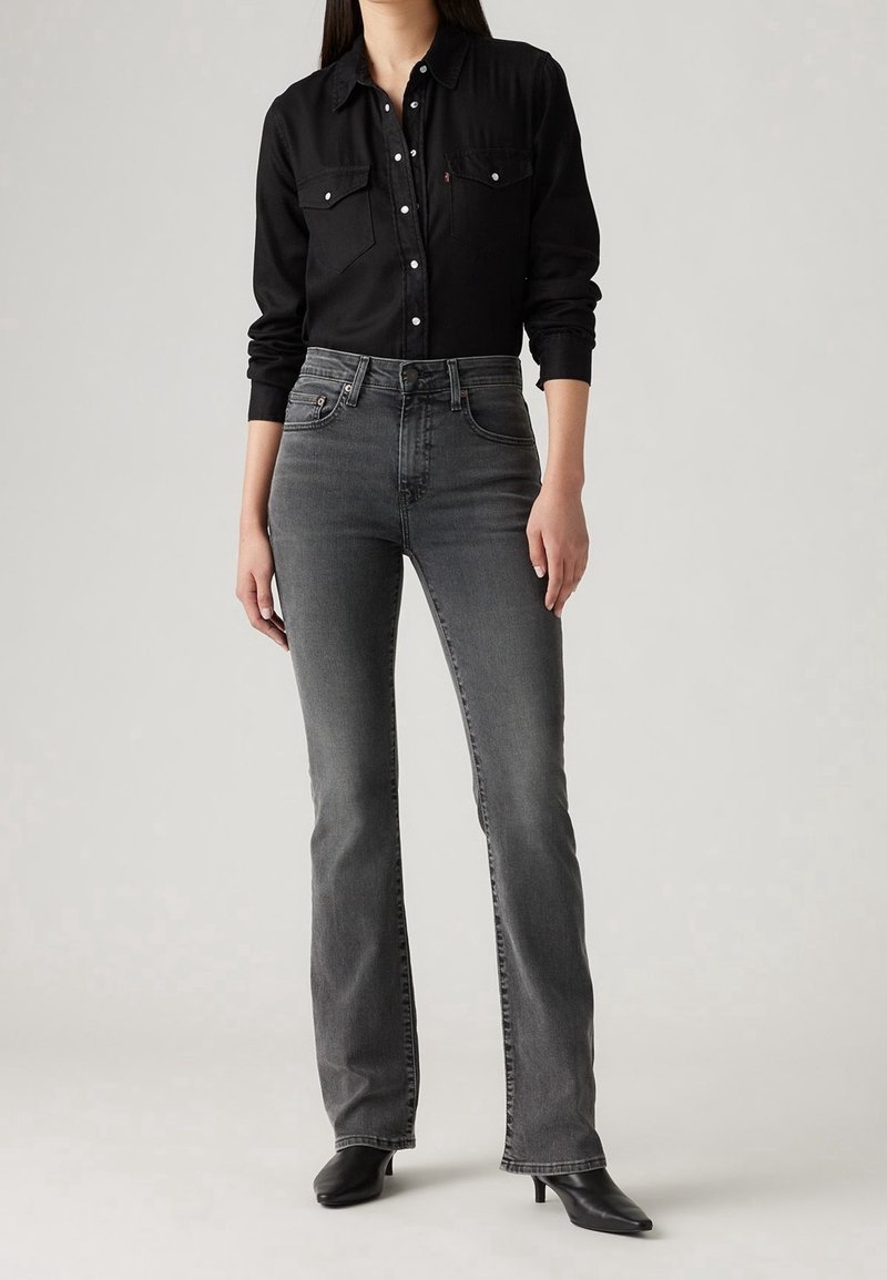 Black button-up shirt with two chest pockets, paired with high-waisted, straight-leg, dark grey jeans. Black heeled ankle boots complete the look.