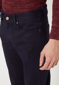 Los pantalones de algodón azul marino presentan un diseño de frente plano y cinco bolsillos, con remaches en tono cobre en la cintura y los bolsillos. La textura parece suave.