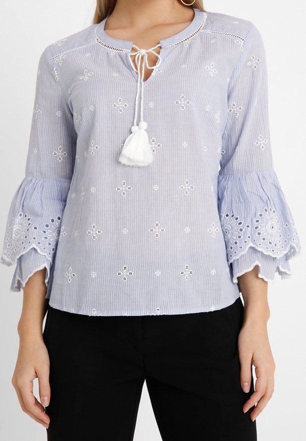 Blouse bleu clair avec motif floral brodé, manches cloche proéminentes, encolure ronde et détail de lien à pompons à l'avant.