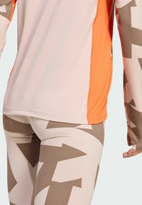 Top met lange mouwen in lichtroze met oranje accenten en platte naden, gecombineerd met een high-waist legging met een bruin pijlenpatroon.