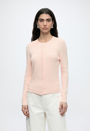 Vrouw met lang donker haar draagt een lichtroze kabelgebreide cardigan met knopen en witte broek, staand tegen een effen lichte achtergrond.