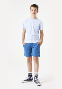 T-shirt en coton bleu clair, shorts bleus et baskets Converse marine avec des accents blancs. Modèle debout les mains dans les poches.