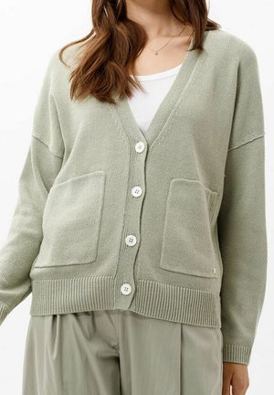 Femme portant un cardigan vert clair boutonné avec deux poches devant sur un top blanc et un pantalon ample vert clair.