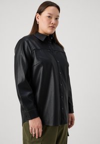 Črna umetna usnjena srajca, z gumbi in ovratnikom ter ramenskimi šivi, ki ima horizontalni šiv čez prsi in dolge rokave.