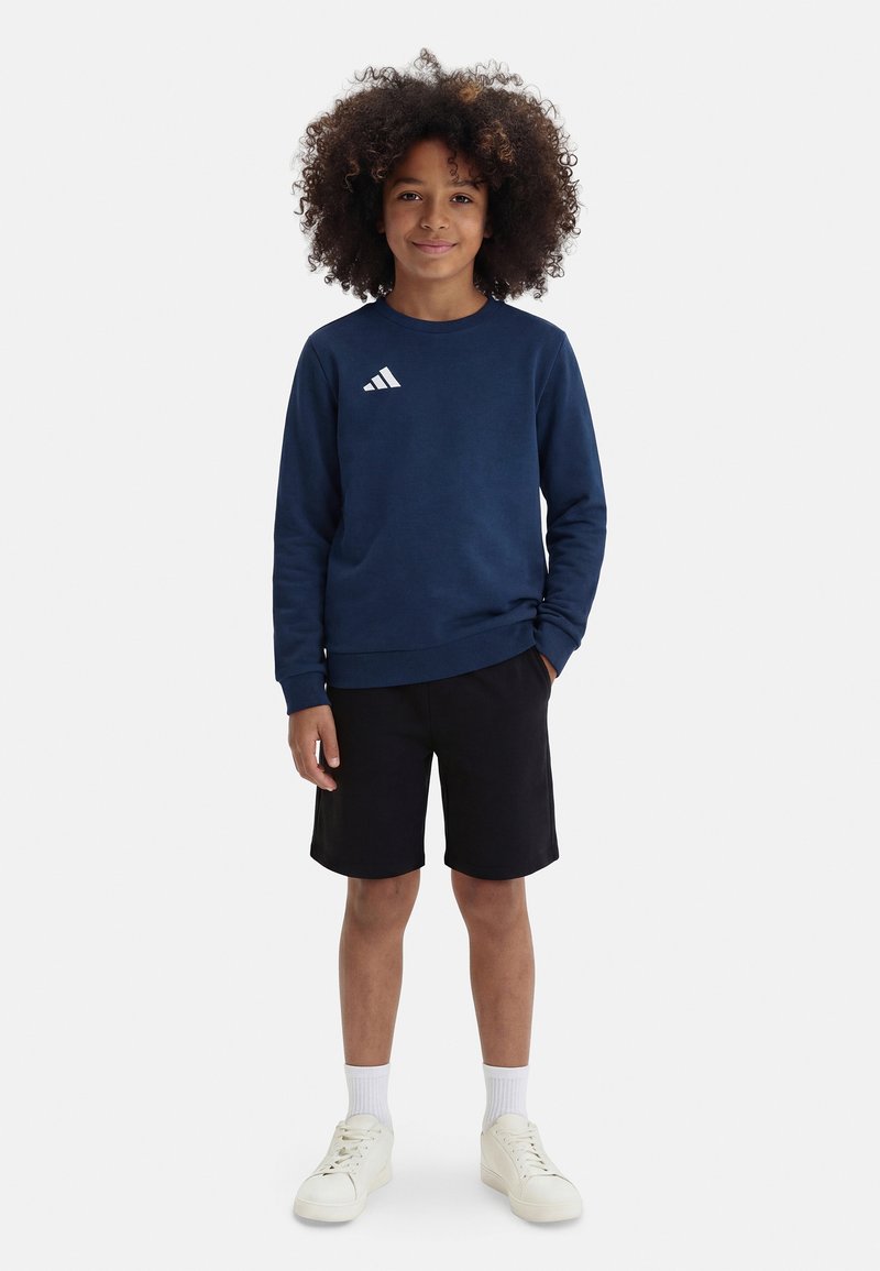 Παιδί με σγουρά μαλλιά που φοράει μπλε σκουρόχρωμο φούτερ, μαύρο σορτς, λευκές κάλτσες και λευκά αθλητικά παπούτσια, στέκεται με το ένα χέρι στην τσέπη.