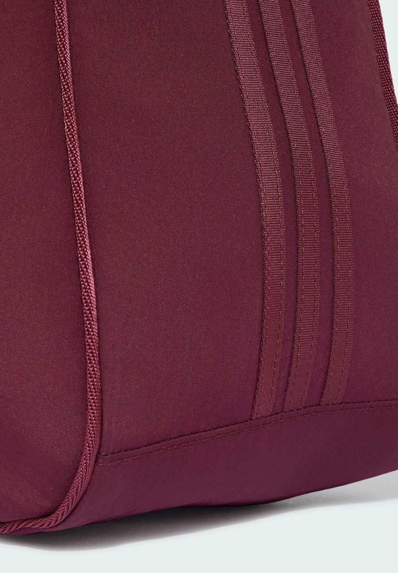 Primo piano dell'angolo di una borsa in tessuto bordeaux con cuciture ricamate e tre strisce verticali strutturate su una superficie liscia.