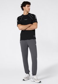 Schwarzes Sport-T-Shirt mit weißen Akzenten und Logo, kombiniert mit grauen Jogginghosen. Model trägt hellgraue Sneaker. Schlichtes, sportliches Design.