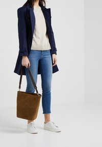 Brun mocka bucketväska med en brun läderrem som bärs av en person i en marinblå kappa och ljus tröja, tillsammans med blå jeans och vita sneakers.