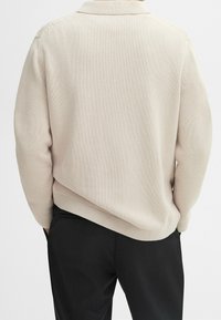 Jasnobeżowy sweter-kardigan z dzianiny z kołnierzem na zakładkę, teksturowaną tkaniną i luźnym krojem, w połączeniu z czarnymi spodniami. Widok od tyłu, styl luźny.