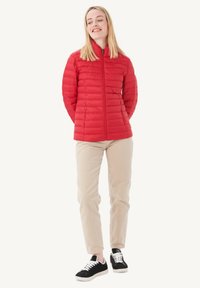 Giacca leggera in piuma rossa con zip frontale, colletto alto e dettagli a cucitura orizzontale. Abbinata a pantaloni beige e sneakers nere.