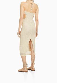 Vestido de ganchillo beige hasta la rodilla con cortes laterales, cuello halter y abertura en la espalda. Llevado con sandalias doradas de tiras.