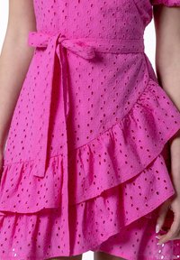 Pink cotton dress featuring a textured eyelet pattern, a fitted waist with a tie belt, and layered ruffles at the hem.