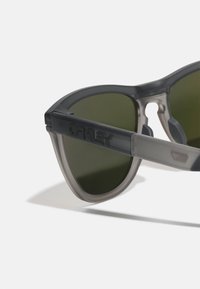Occhiali da sole neri e grigi con montatura sfumata, lenti verde scuro e logo inciso sul lato. Presentano un design elegante e affusolato.