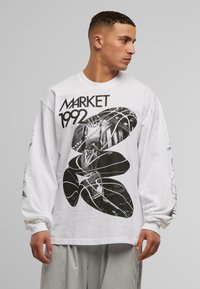 Fehér, hosszú ujjú póló, amelynek feltűnő fekete grafikai tervezése van, rajta a "MARKET 1992" felirattal és absztrakt formákkal. Laza szabás, puha pamut anyag.