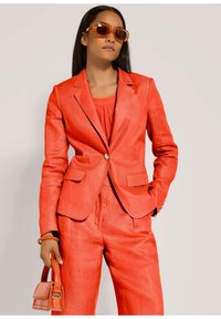 Rode linnen blazer met bijpassende broek, voorzien van een enkele gouden knoop, op maat gemaakt en met zakken. Accessoires zijn een oranje handtas.