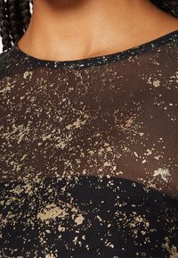 Durchsichtige schwarze Bluse mit goldenen Spritzmustern, runder Ausschnitt und strukturierten Finish, das sich mit einem soliden schwarzen Abschnitt kontrastiert.