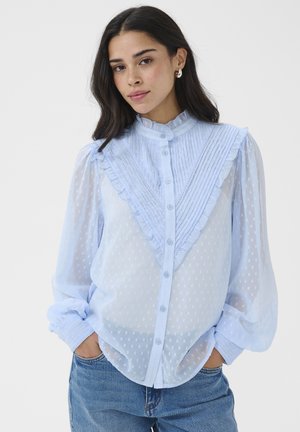 Mladá žena v priesvitnej svetlomodrej blúzke s volánikmi a textúrovaným vzorom, ktorá má na sebe modré džínsy, pózuje s rukami v kapse na bielom pozadí.