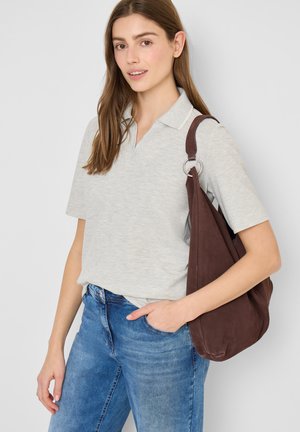 Giovane donna con polo a maniche corte grigio chiaro, jeans blu e borsa marrone a tracolla, in posa contro uno sfondo chiaro e uniforme.