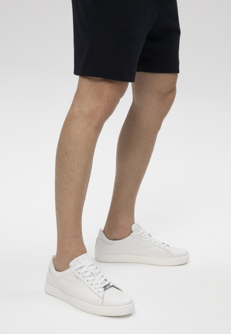 Personne portant un short noir et des baskets blanches basses debout sur un sol blanc.