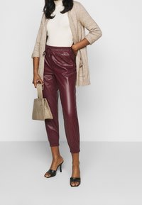 Burgundy konstläder joggers med elastiska muddar, kombinerat med en beige stickad cardigan och svarta öppen-tå skor, håller en strukturerad taupe handbag. 