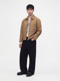 Jeune homme portant une veste zippée beige, un t-shirt graphique, un pantalon large noir et des chaussures noires, debout devant un fond blanc uni.