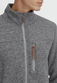 Giacca grigia con zipper e colletto alto, in tessuto texturizzato e con tasca sul petto. Presenta una cerniera chiara in contrasto e un accento in corda marrone.