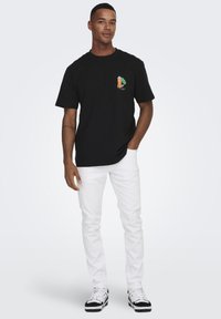 Black cotton t-shirt featuring a colorful cartoon graphic on the left chest, paired with white pants and black and white sneakers.