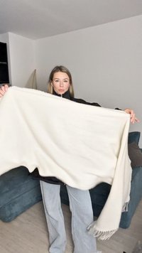 Pătură dintr-un material moale, alb, cu o textură cu nervuri și margini ciufulite, ținută de o persoană îmbrăcată într-o geacă neagră și blugi deschiși.