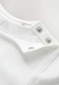 Hvid babybeklædningshalsudskæring med to metaltrykknapper delvist åbne på blødt bomuldsstoffet.