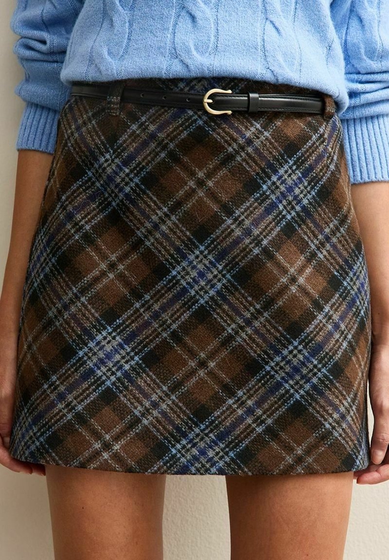 Pull bleu torsadé rentré dans une jupe en laine à carreaux marron avec ceinture noire et boucle dorée, porté par une personne debout.