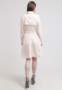 Jasnoróżowy płaszcz typu trencz z paskiem, z warstwowym tyłem i długimi rękawami. Miękki, gładki materiał o minimalnym połysku.