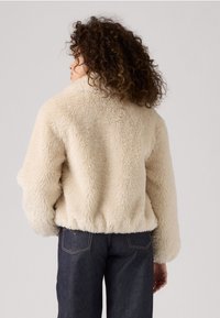 Crème-kleurige nepbontjas met een zachte, gestructureerde afwerking; cropped ontwerp, hoge kraag en elastische tailleband. Draagt bij donkere denim broeken.