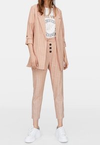 Blazer rayé rose clair avec pantalon assorti et ajusté. Stylisé avec un t-shirt graphique blanc et des baskets blanches. Comprend des détails à boutons.