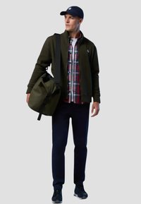 Giacca verde oliva, camicia a quadri, pantaloni blu navy e sneakers, con una grande borsa a tracolla verde oliva. Un cappello scuro con logo completa il look.