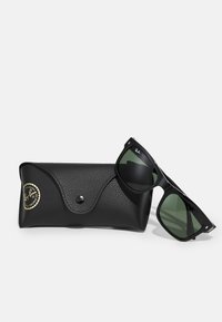 Ochelari de soare negri Ray-Ban cu lentile verde închis și ramă unghiulară, alături de un toc din piele texturată neagră, cu un logo auriu.