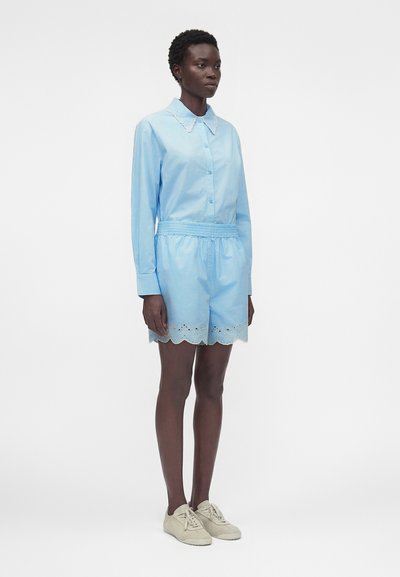 Γυναίκα που στέκεται, φοράει ανοιχτό μπλε πουκάμισο με κουμπιά και κυματιστό γιακά και ταιριαστό σορτς με κυματιστό τελείωμα, μπεζ αθλητικά παπούτσια, ουδέτερο φόντο.