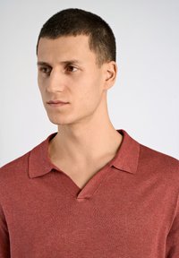 Röd stickad polotröja med V-ringning och kort krage. Tyget verkar mjukt, med en subtil textur och jämn färg genom hela plagget.