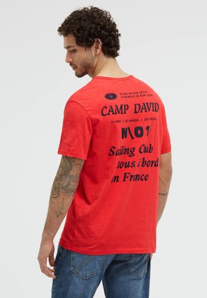 Man with curly hair and tattooed arm wearing red Camp David T-shirt and blue jeans, turned slightly to show back text.