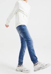Sudadera blanca con capucha, jeans ajustados azul y zapatillas gris. Los jeans presentan un corte clásico con bolsillos traseros y detalles desgastados.