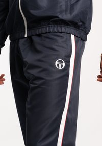 Pantaloni da allenamento navy in materiale leggero, con una striscia laterale bianca e rossa, vita elasticizzata e logo bianco sulla coscia.