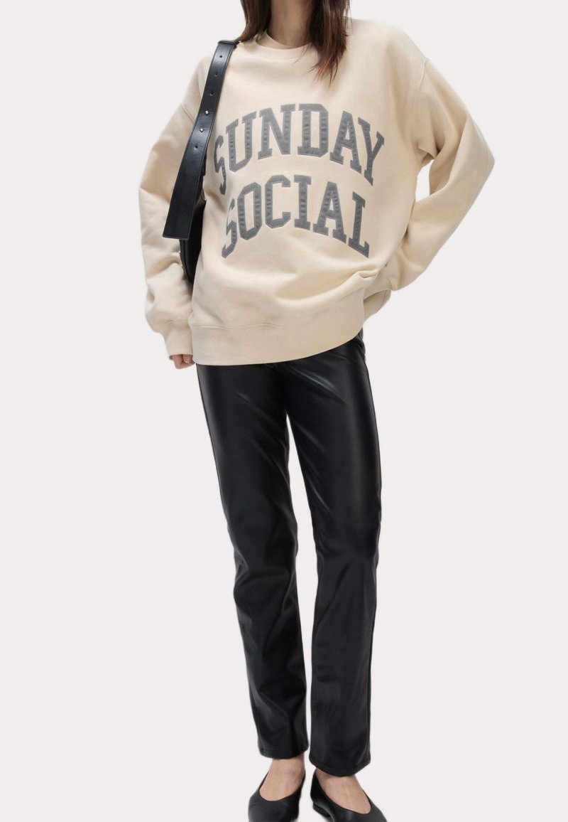 Femme portant un sweat-shirt beige avec le texte "SUNDAY SOCIAL", un pantalon en cuir synthétique noir, des ballerines noires et un sac à bandoulière noir.
