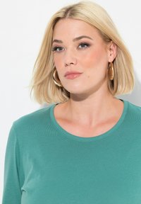 Smaragdgrünes Langarmshirt aus weichem Stoff mit runder Ausschnitt und sichtbaren Texturdetails, kombiniert mit großen goldenen Creolen.