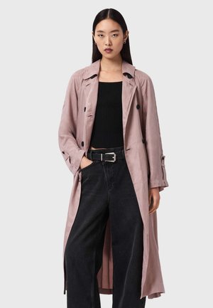 Hellrosa Trenchcoat mit glatter Textur, doppelreihigem Design und schwarzen Knöpfen. Getragen über einem schwarzen, gerippten Oberteil und schwarzen Jeans.