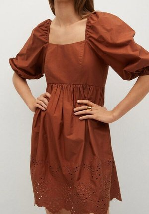 Kobieta w rudej sukience z bufiastymi rękawami, kwadratowym dekoltem i florystycznym wykończeniem z haftem, z rękami na biodrach, nosząca dwa złote pierścionki na prawej ręce.