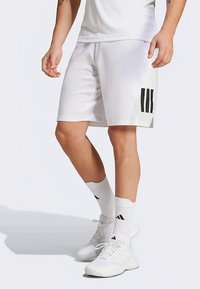 Persona valkā baltas sporta biksītes ar melnām svītrām, baltas pusmitras zeķes un baltus sporta apavus, stāvot pret vienkāršu fona. 