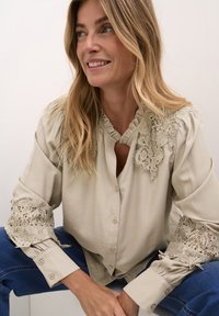 Blouse beige avec col à volants et accents en dentelle délicate sur les manches. Corps uni, fermeture à boutons, manches longues et coupe décontractée.