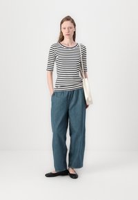 Gestreiftes, eng anliegendes Top in Schwarz und Weiß mit 3/4-Ärmeln; lockere blaue Denim-Hose; schwarze flache Schuhe; cremefarbene Umhängetasche; glatte Textur.
